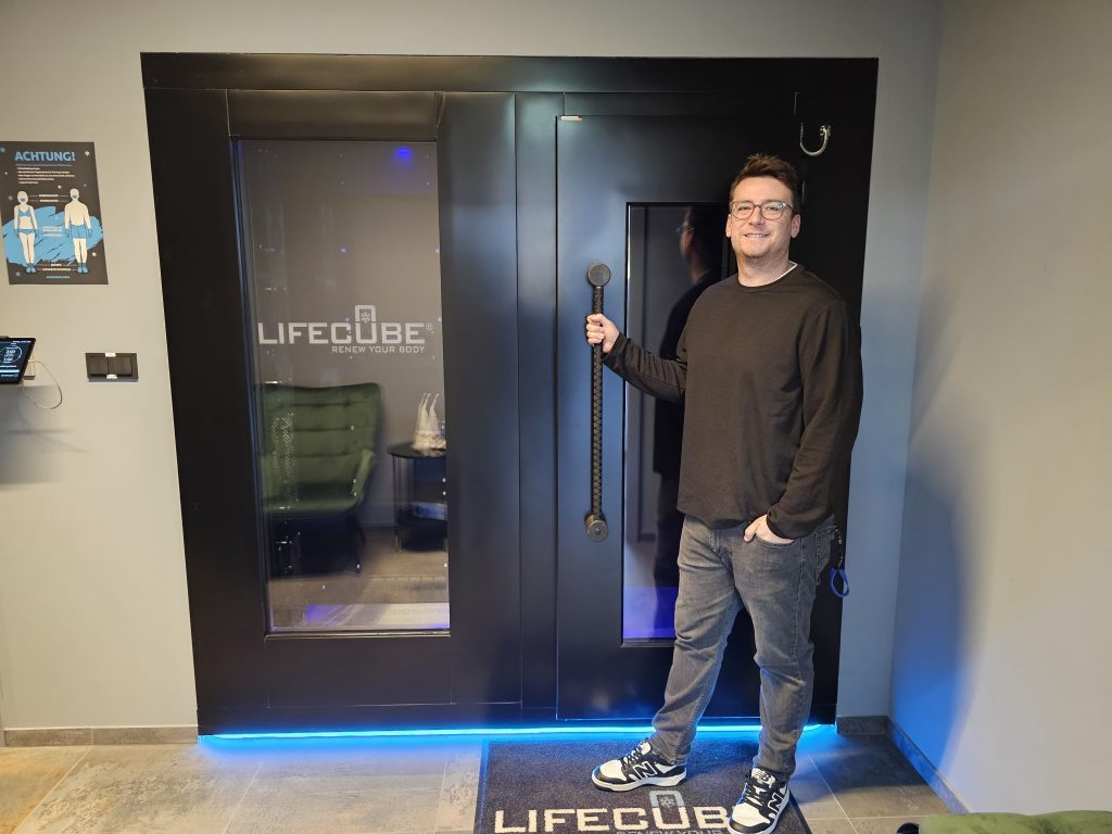 Ein lächelnder Mann mit Brille hält den Griff einer schwarzen LifeCube-Kabine mit blauem Bodenlicht in einem Fitnessraum.