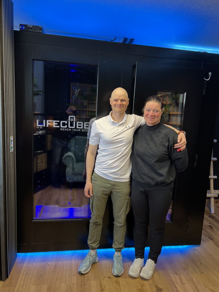Ein lächelndes Paar posiert Arm in Arm vor einer schwarzen LifeCube-Kabine mit auffälliger blauer Beleuchtung.