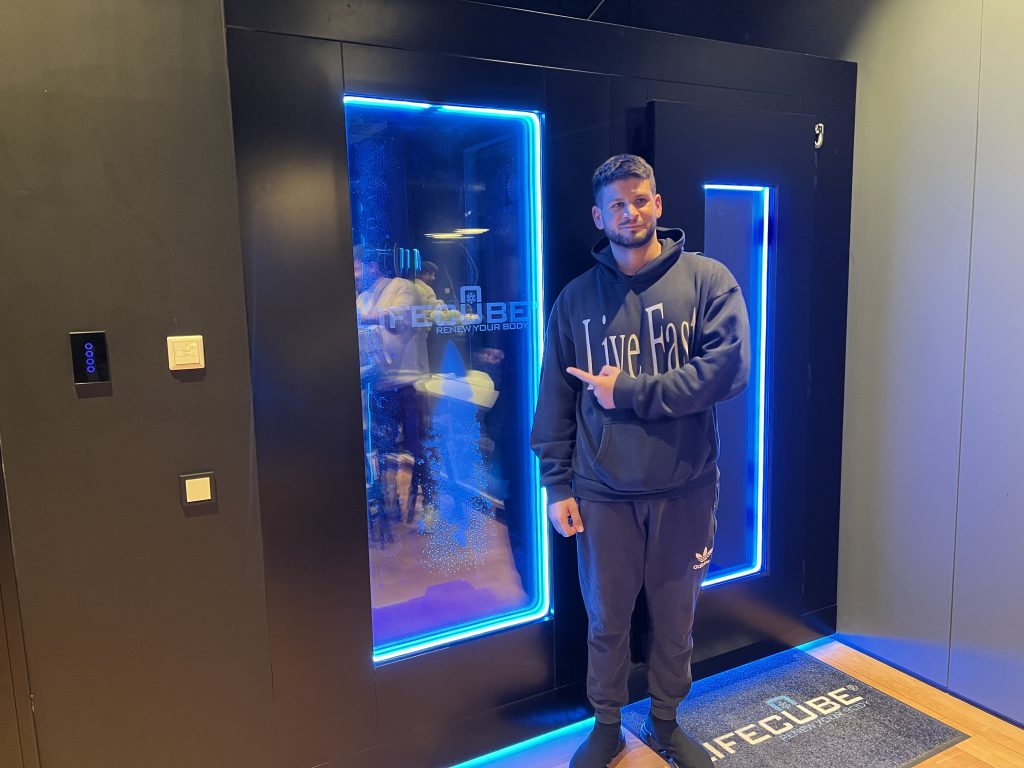 Ein junger Mann im dunklen Kapuzenpullover zeigt auf die blau beleuchtete Glastür einer LifeCube-Anwendungskabine.