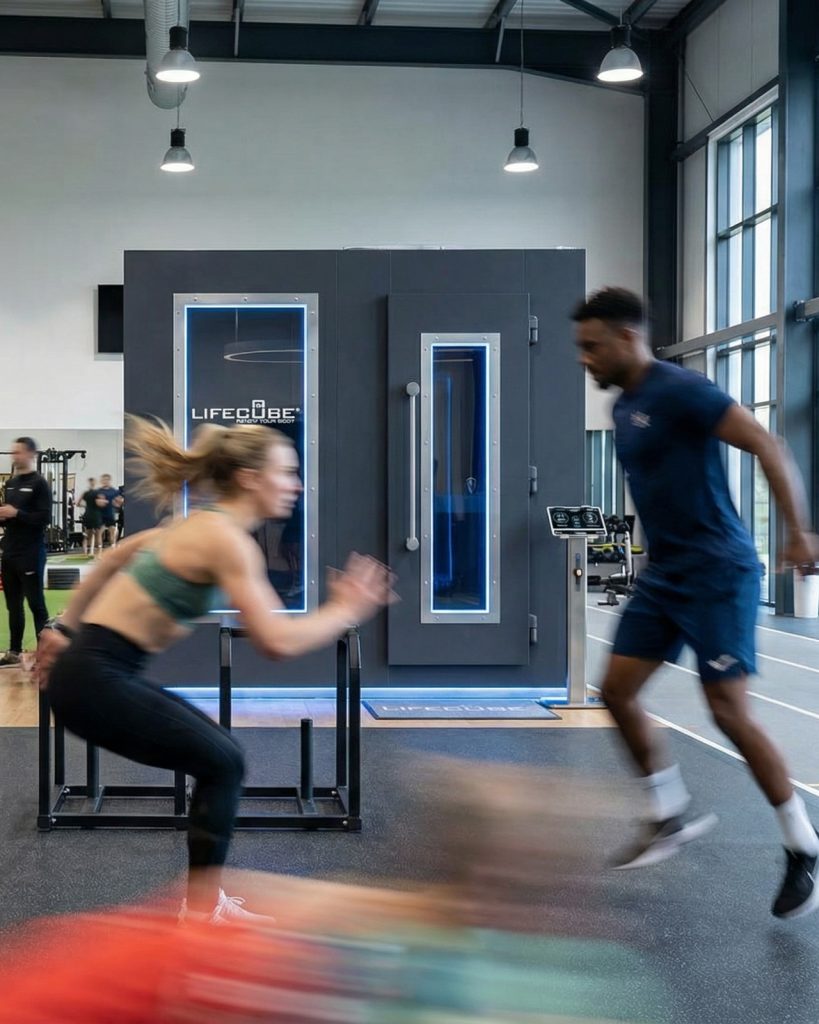 Zwei Sportler trainieren dynamisch vor einer dunklen LifeCube elektrische Kältekammer in einem modernen Fitnessstudio.
